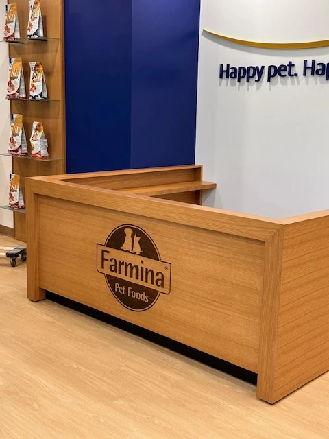 A custom oak reception desk with a laser-etched Farmina Pet Foods logo, set against a blue and white wall with the "Happy pet. Happy life." sign, and surrounded by oak floating shelves and wall panels.